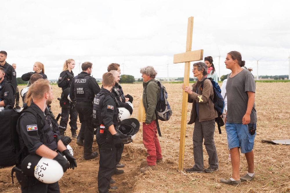 Foto vom 21. Juli 2022 Menschen mit gelbem Kreuz vor einer Gruppe Polizist:innen. Letzere schützen RWE bei der Einwallung von Lützerath