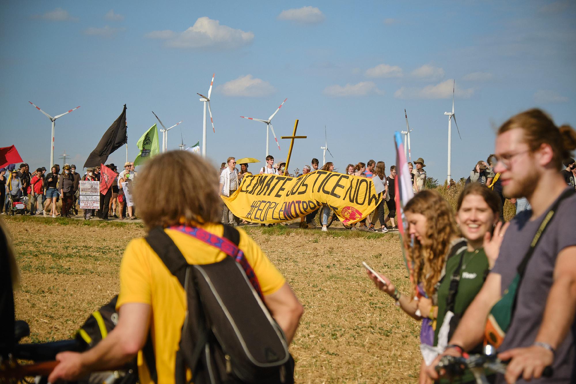 Bild der Demo vom 3. September 2022 mit gelbem Kreuz und Bannern und Fahnen. Menschen gehen von Keyenberg nach Lützerath über einen Feldweg.