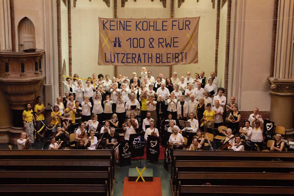 Solifoto des Chors und Orchesters Lebenslaute mit Lützerath. Es zeigt ca 100 Musizierende in der Taborkirche, Kreuzberg unter einem Banner mit der Aufschrift Keine Kohle für A100 und RWE. Lützerath bleibt!