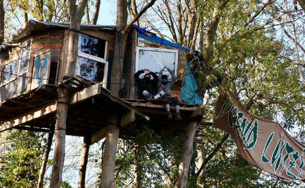 Foto eines Baumhauses mit zwei Menschen, die von der Plattform zu uns runterschauen und ein Herz mit ihren Fingern formen. Rechts im Bild ein Banner mit der Aufschrift Defend Lützi.