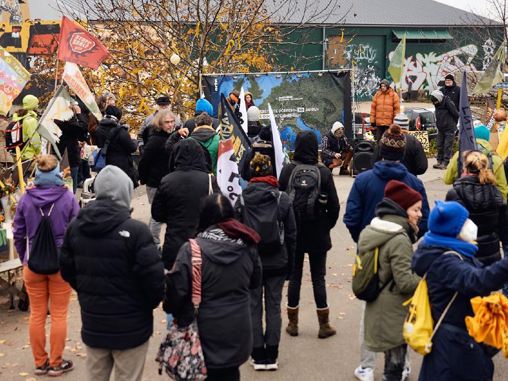 Bild eines November Dorfspaziergangs. Eine Gruppe von Menschen schaut auf eine Karte Europas, worauf steht, wieviele Dörfer in Deutschland schon zerstört, wieviele und Menschen wegen Braunkohle umgesiedelt wurden. Im Hintergrund die Skatehalle mit Wandgemälden von Captain Borderline.