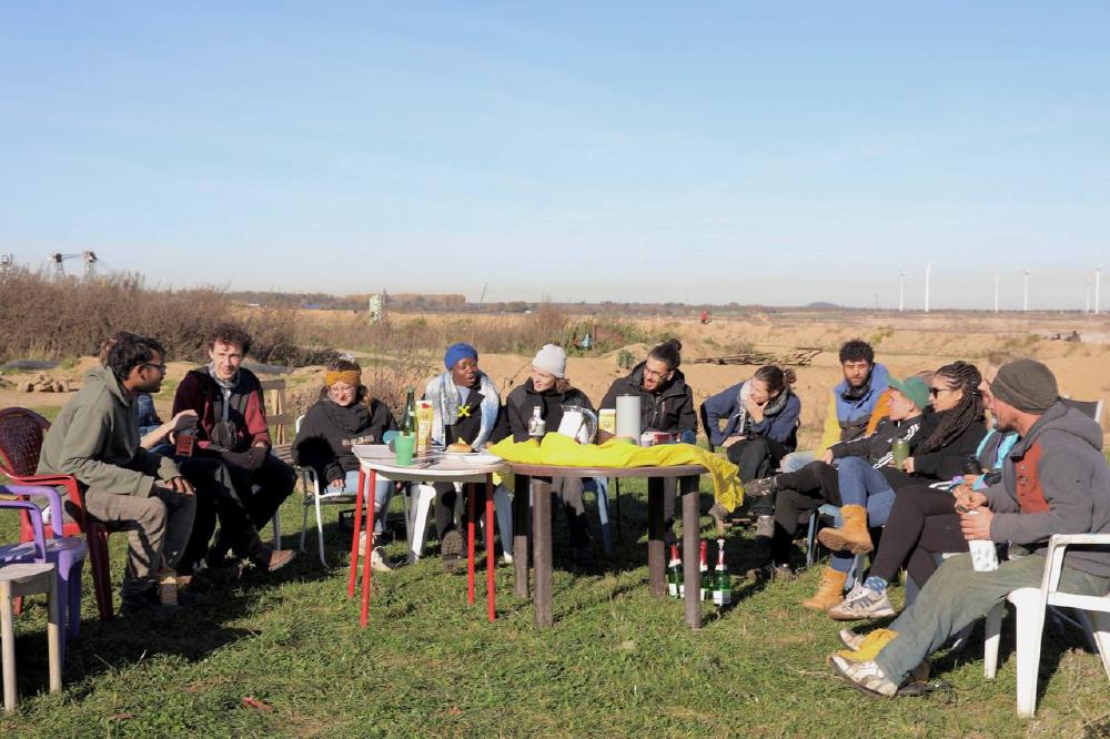 Foto der Pressekonferenz mit Kaossara und weiteren sieben Menschen, die an einem langen Tisch an der Kante beim Wendehammer sitzen. Im Hintergrund der Tagebau Garzweiler, links ist ein Schaufelradbagger zu erkennen.