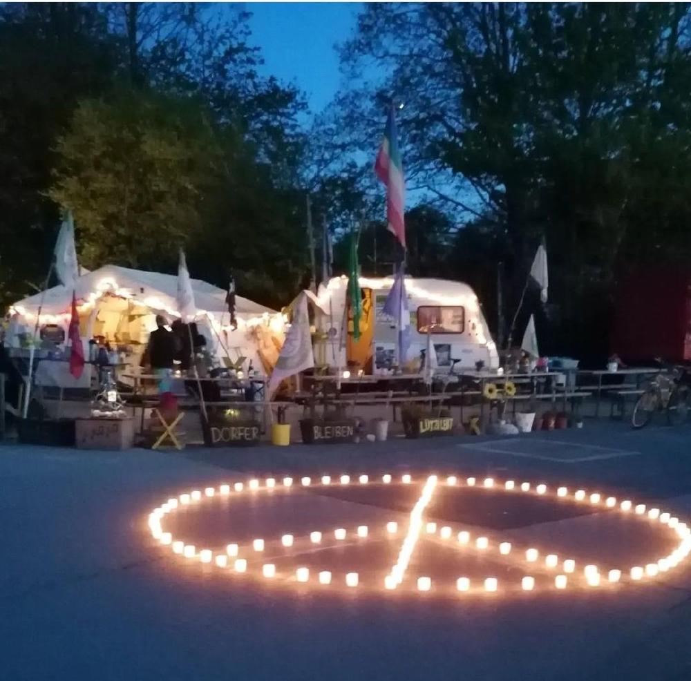 Bild zeigt die Mahnwache Lützerath, ein große Zelt, ein Wohnwagen, davor sind Tische und Bänke, so wie ganz viele Fahnen. Unten auf dem Bild vor der Mahnwache ist aus Kerzen das Peace Symbol zu sehen.