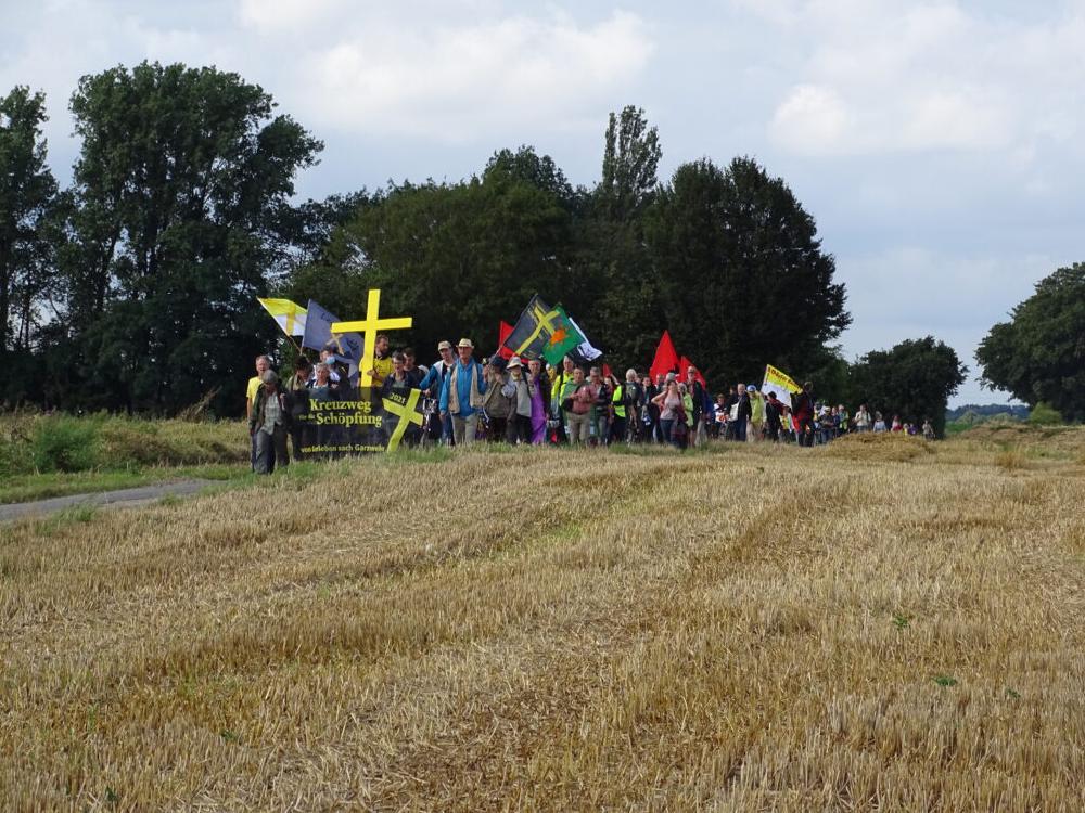 Ein Bild des Kreuzwegs 2021 von Gorleben nach Lützerath. Eine Menschengruppe, an der Spitze das gelbe Kreuz und ein Banner mit der Aufschrift Kreuzweg für die Schöpfung 2021.