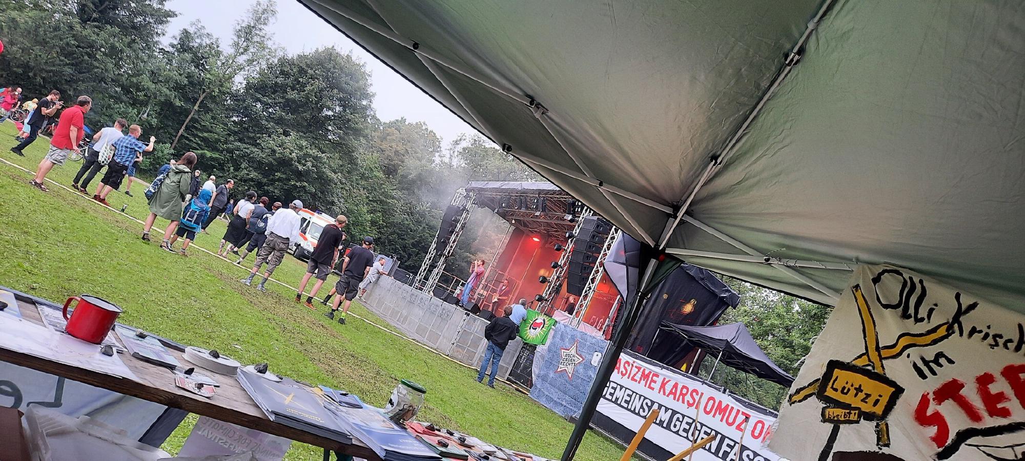 Bild der Veranstaltung mit Sicht auf die Bühne, im Vordergrund ein Tisch mit Infomaterial und Banner auf denen Gemeinsam gegen den Faschismus in zwei Sprachen und Lützi bleibt! zu lesen sind