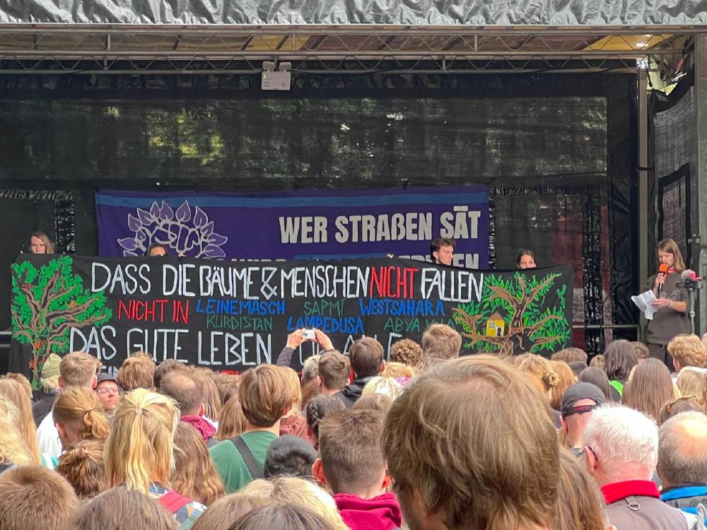 Banner der Leinemaschdemo vom 1. Oktober 2023 mit dem Schriftzug Dass die Bäume und Menschen nicht fallen. Nicht in Leinemasch, Sapmi, Westsahara, Kurdistan, Lampedusa, Abya Yala. Das Gute Leben für Alle.